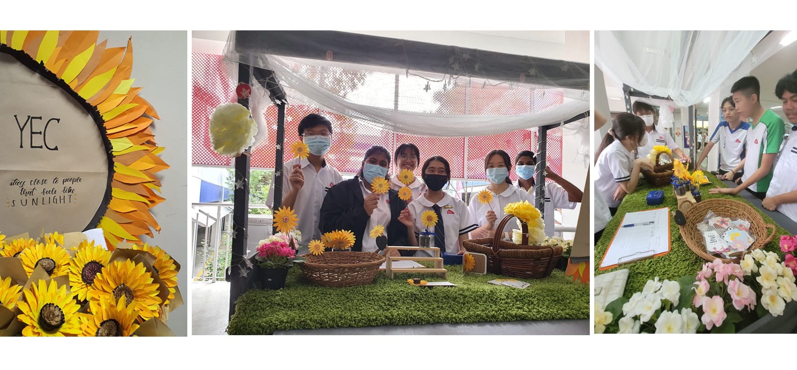Students in masks hold paper sunflowers at a decorated booth with baskets and artificial grass at a school event.